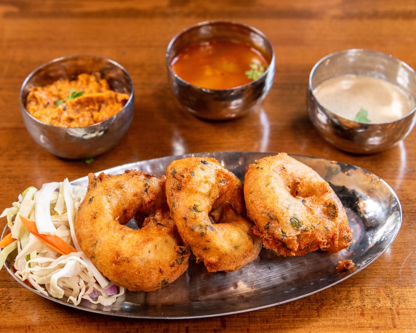Three golden-fried vada donuts with shredded cabbage salad and three small bowls of dipping sauces.