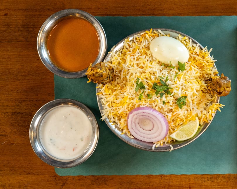 Chicken biryani with a boiled egg, onion, lime, and two side sauces on a green mat.