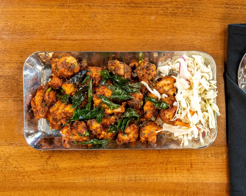 A metal platter of fried chicken pieces with curry leaves and a side of shredded cabbage.