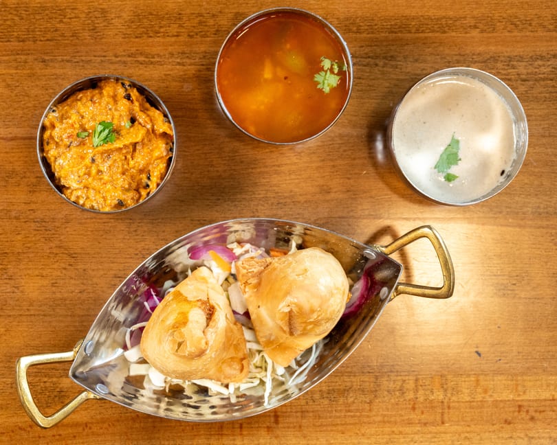 Two golden-brown samosas in a silver dish with shredded cabbage and red onions, alongside three bowls of sauces.