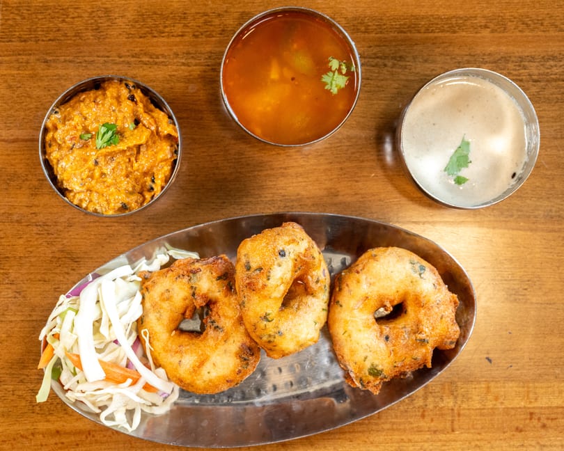 Three golden-brown medu vada, shredded cabbage salad, and two bowls of chutney on a wooden table.