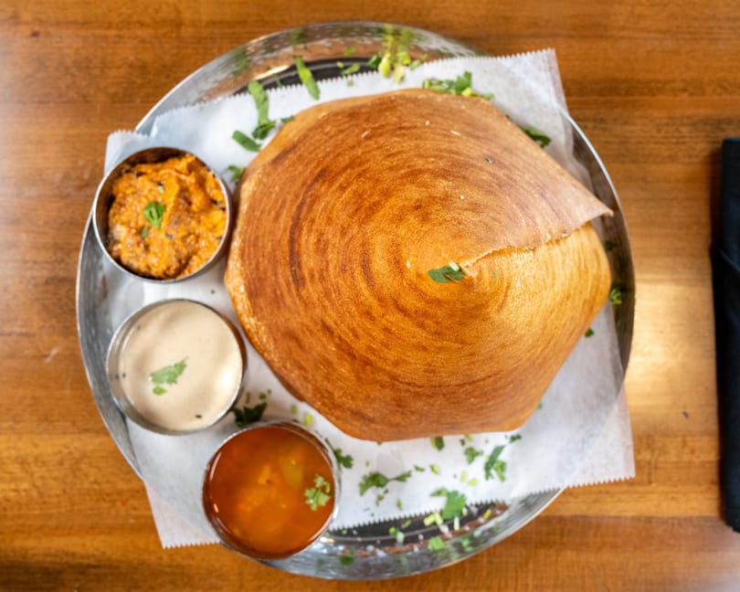 Crispy golden dosa served with three small bowls of colorful chutneys and sambar on a metal plate.
