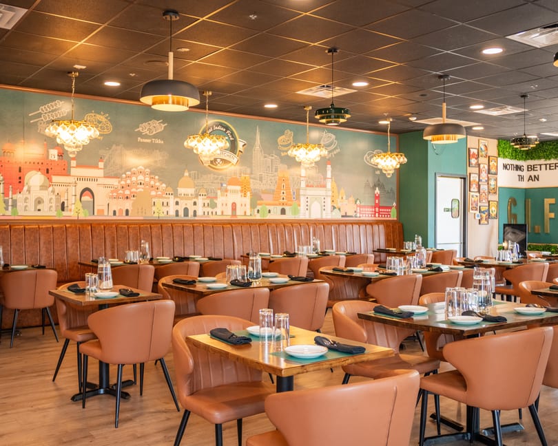 A modern Indian restaurant interior with brown leather seating, wooden tables, and a colorful mural of Indian landmarks.