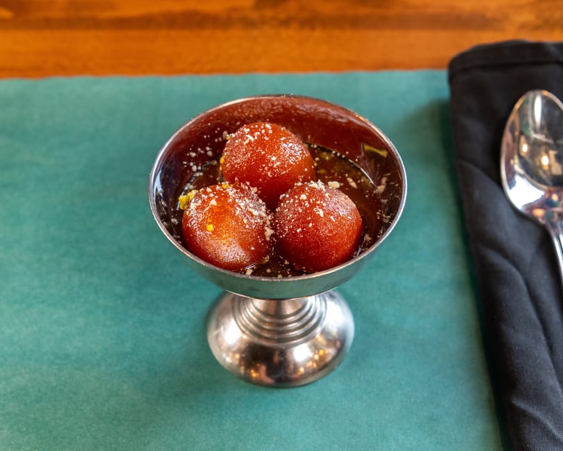 Three gulab jamun in a silver cup, garnished with powdered sugar and pistachios, on a green placemat.