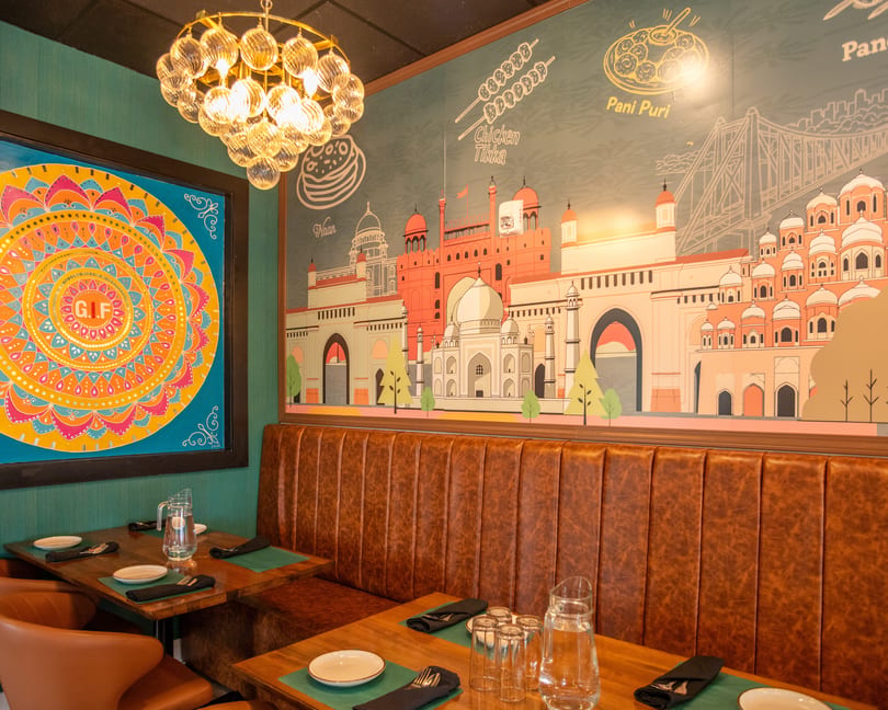 Restaurant interior with brown leather booths, wooden tables, and a colorful mural depicting Indian landmarks and food.